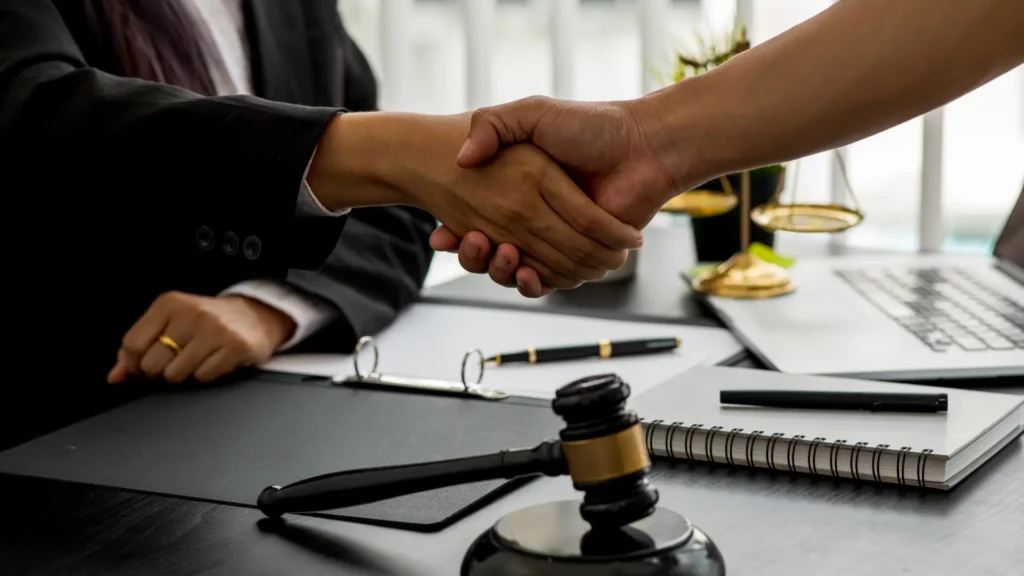 Two individuals shaking hands over a desk with a gavel and legal documents in the foreground.